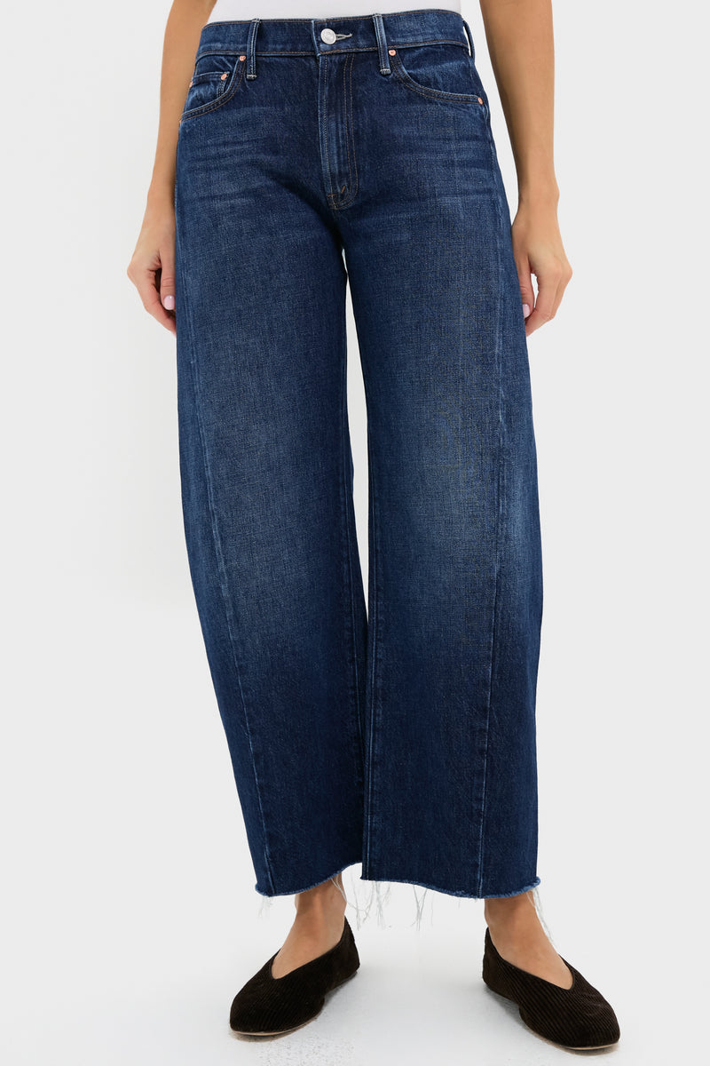 A person wears MOTHER high-waisted, dark blue barrel leg jeans with a frayed hem and black flats, standing in a simple white space.