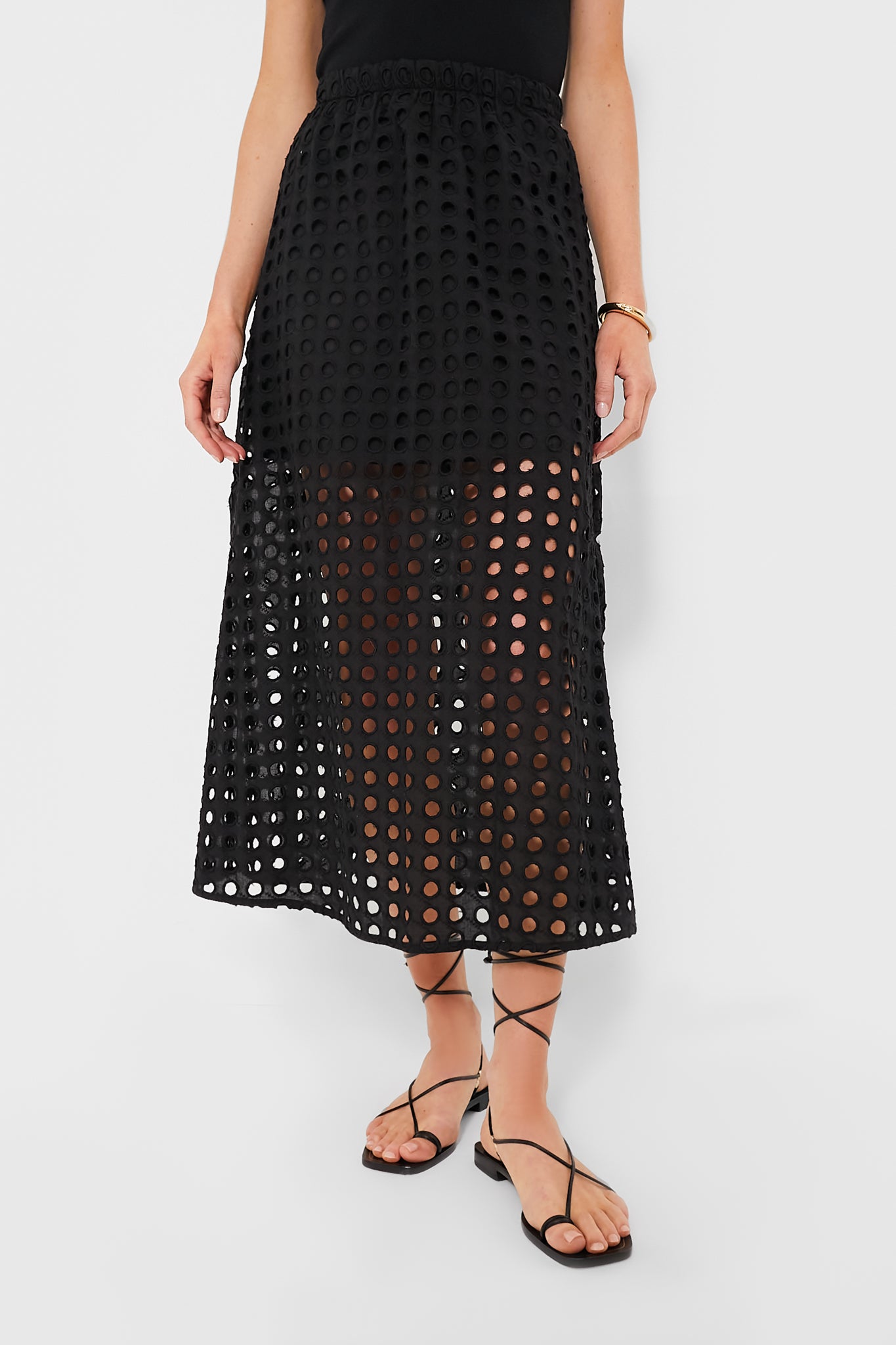 A person wears the Tuckernuck black cotton eyelet skirt with circular cutouts over a mini skirt, featuring an elastic waistband, paired with a black top and strappy black sandals.