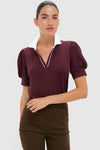 A woman wears a Tnuck Sport burgundy puff-sleeve polo with a white collar and dark brown pants.