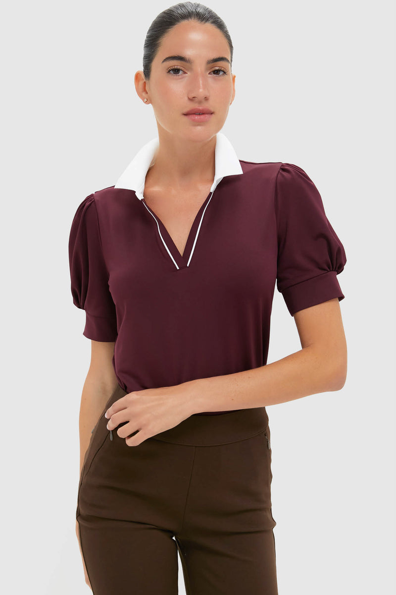 A woman wears a Tnuck Sport burgundy puff-sleeve polo with a white collar and dark brown pants.