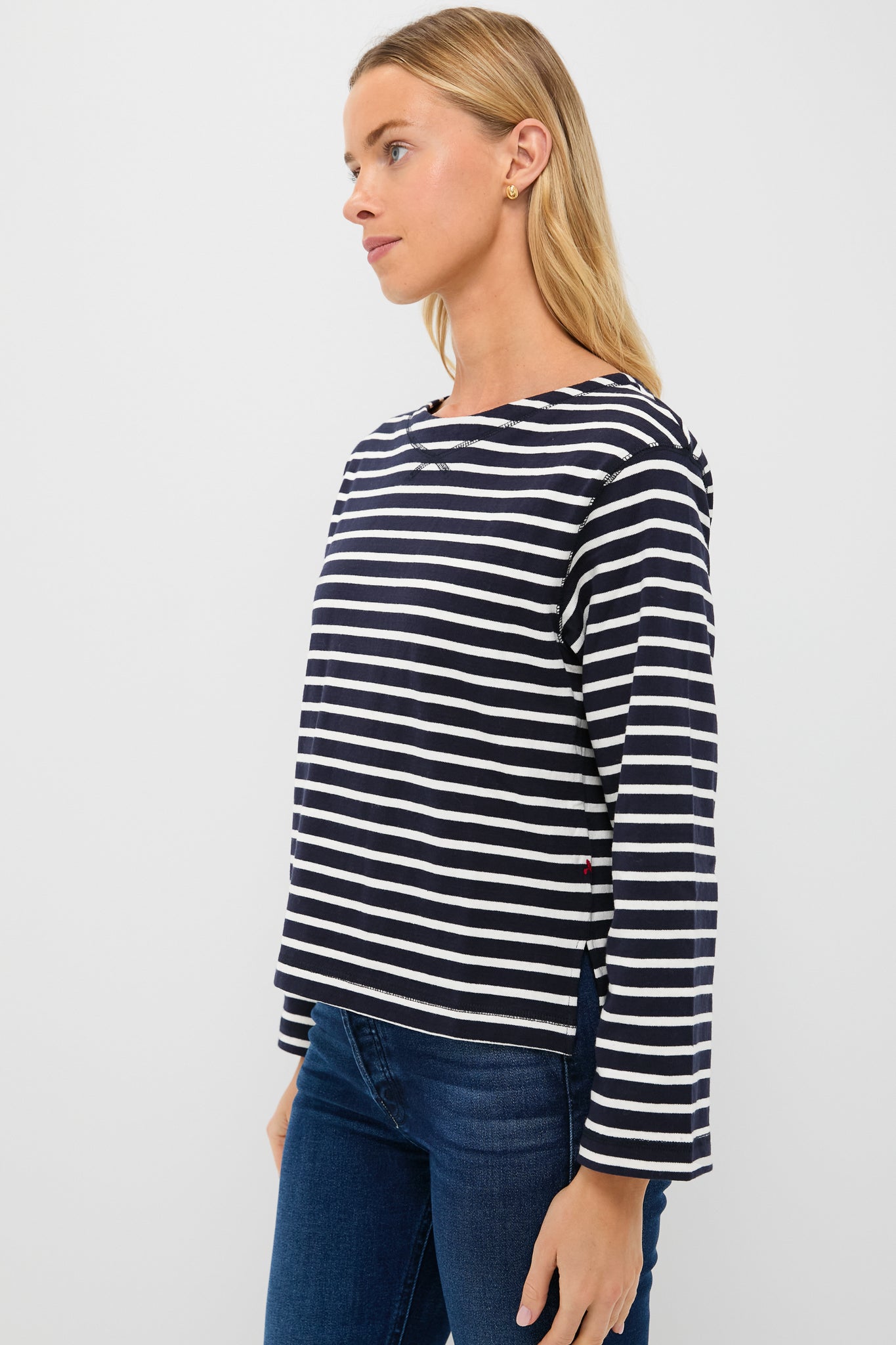 A woman showcases Parisian-chic style in an Alex Mill navy and white striped long sleeve boat neck tee with blue jeans, set against a plain white background.
