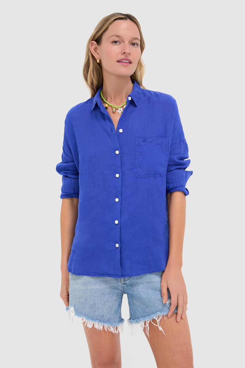 A woman stands confidently in an Alex Mill bright blue linen button-down shirt with rolled sleeves and light denim frayed shorts.