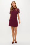 Woman models the classic Oxblood Lace Agatha Mini Dress, featuring an effortless fitted silhouette with sheer lace shoulders, embodying fun, high-end elegance for versatile occasions.
