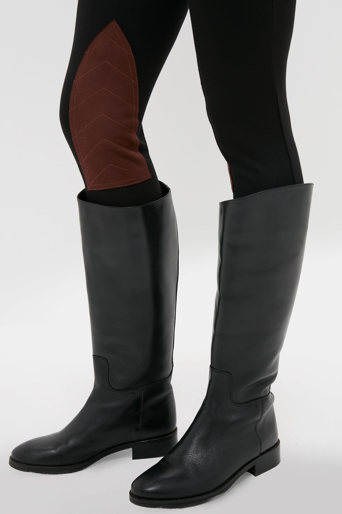 A person wearing Tuckernuck Shoes black knee-high leather boots and black leggings with brown chevron-patterned patches on the inner knees.