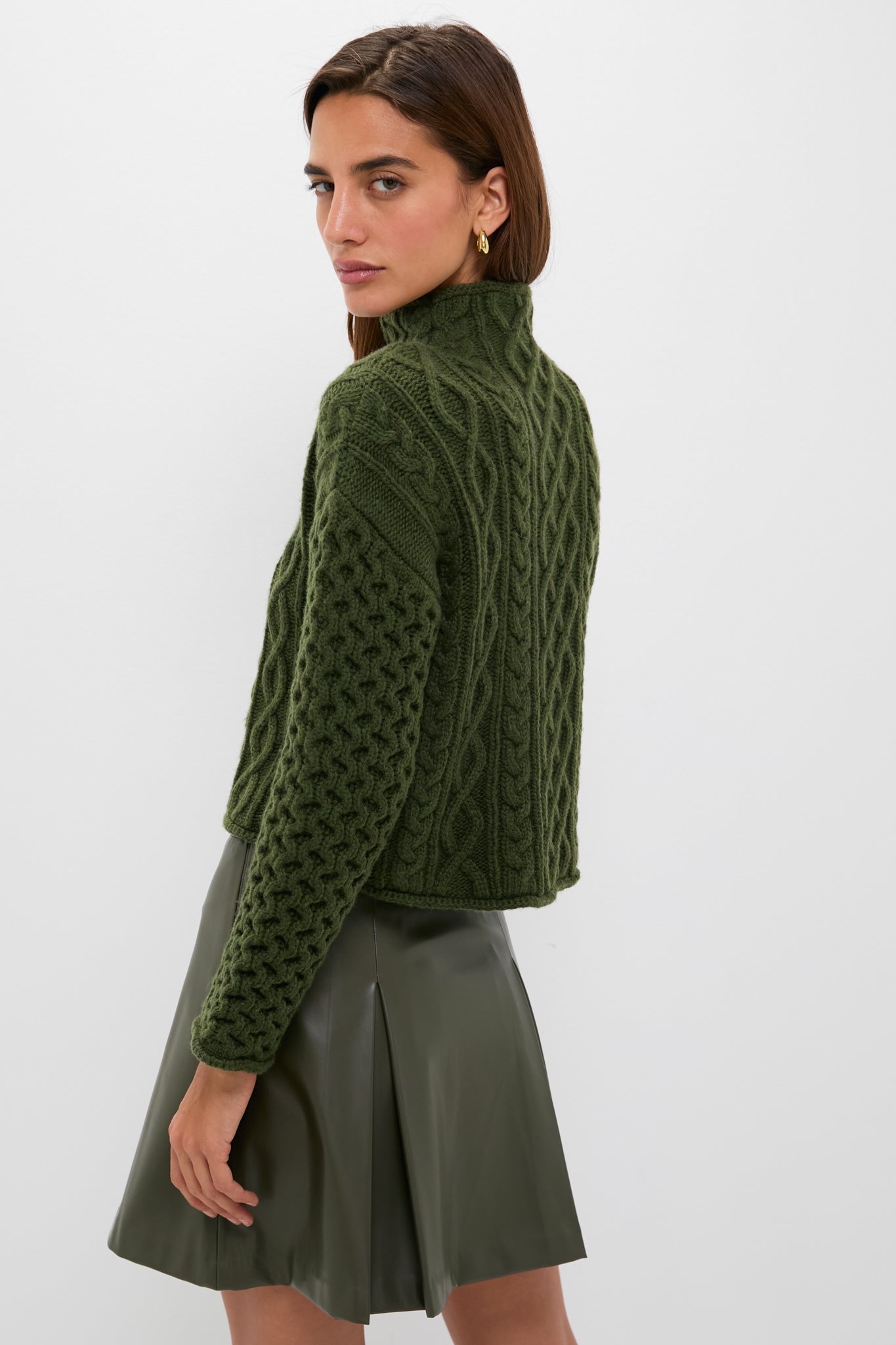Woman in Olive Cable Knit Cropped Perry Turtleneck, showcasing classic, effortless style with a chunky design, paired with a high-end, fun, pleated faux leather skirt.