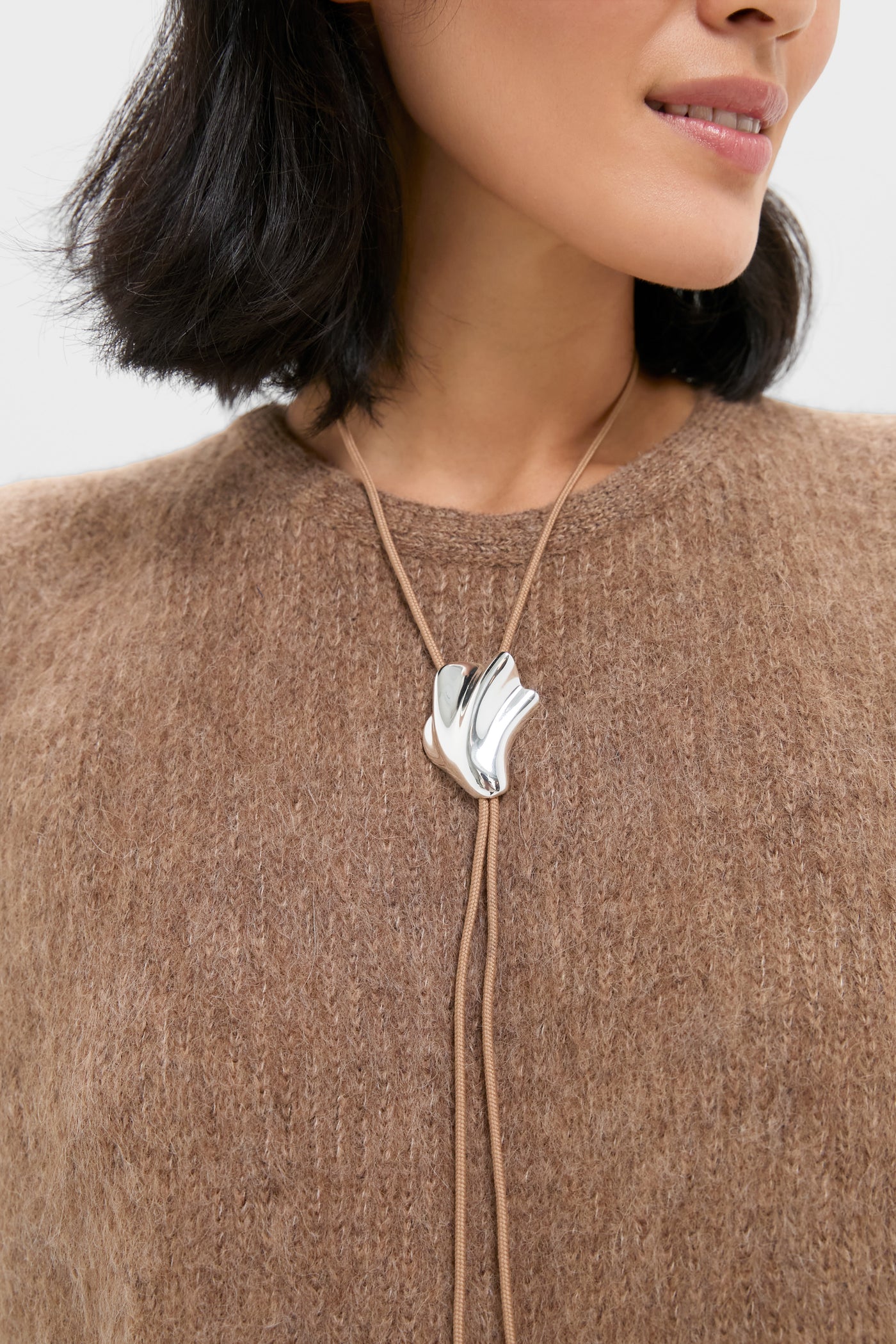 Close-up of a person wearing the High Polish Silver Lydia Bolo Tie Necklace, featuring a sleek, abstract silver pendant on a taupe cord, for an effortless, high-end look.