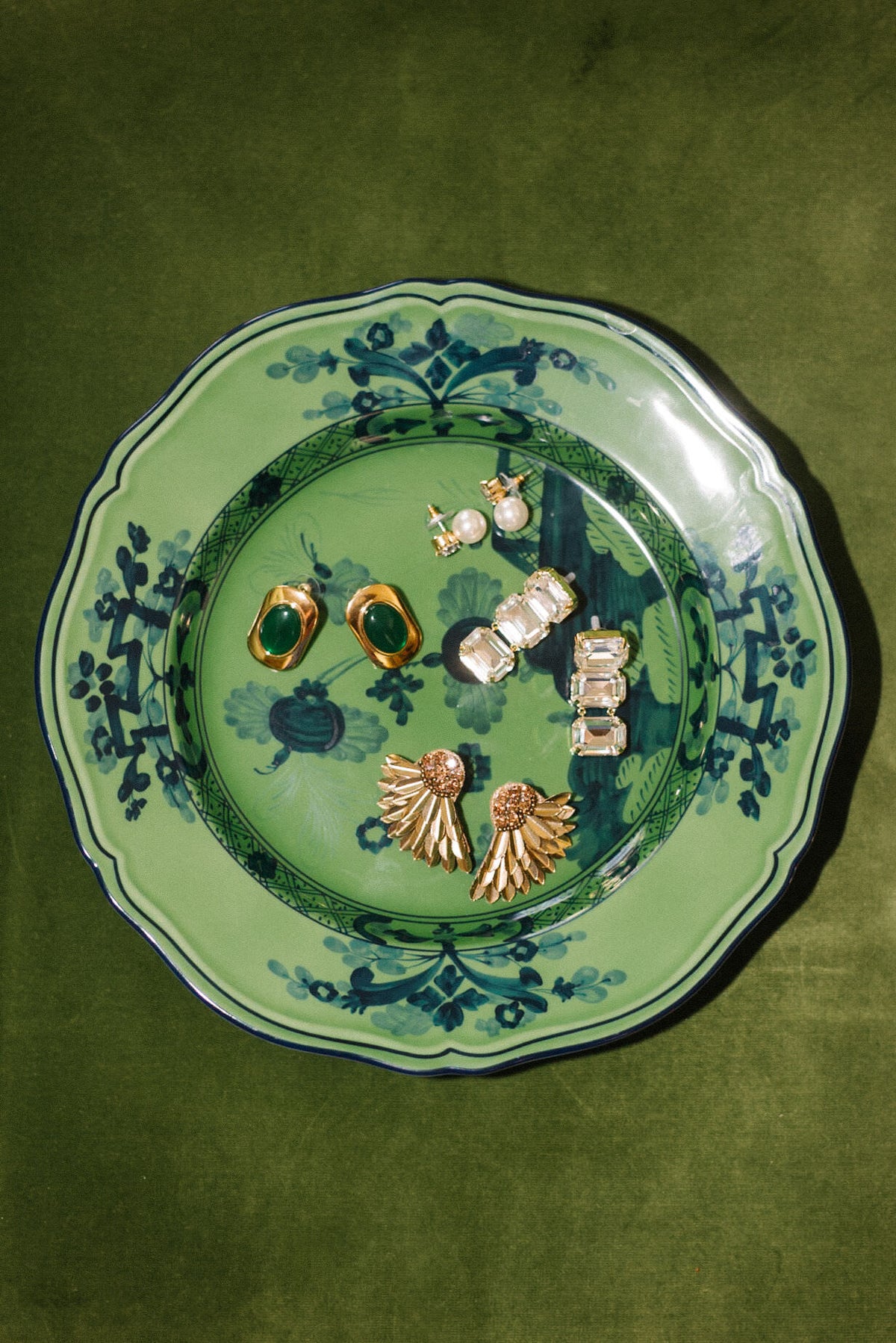 A Ginori 1735 Oriente Italiano Malachite green porcelain salad plate showcases six pairs of gold and gemstone earrings arranged elegantly on a green velvet surface.