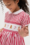 Young girl wearing the Candy Cane Stripe Smocked Dress with classic Peter Pan collar, puff sleeves, and festive gingerbread and candy cane embroidery. Effortless holiday charm in high-end style.
