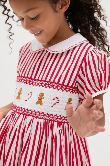 Young girl wearing the Candy Cane Stripe Smocked Dress with classic Peter Pan collar, puff sleeves, and festive gingerbread and candy cane embroidery. Effortless holiday charm in high-end style.