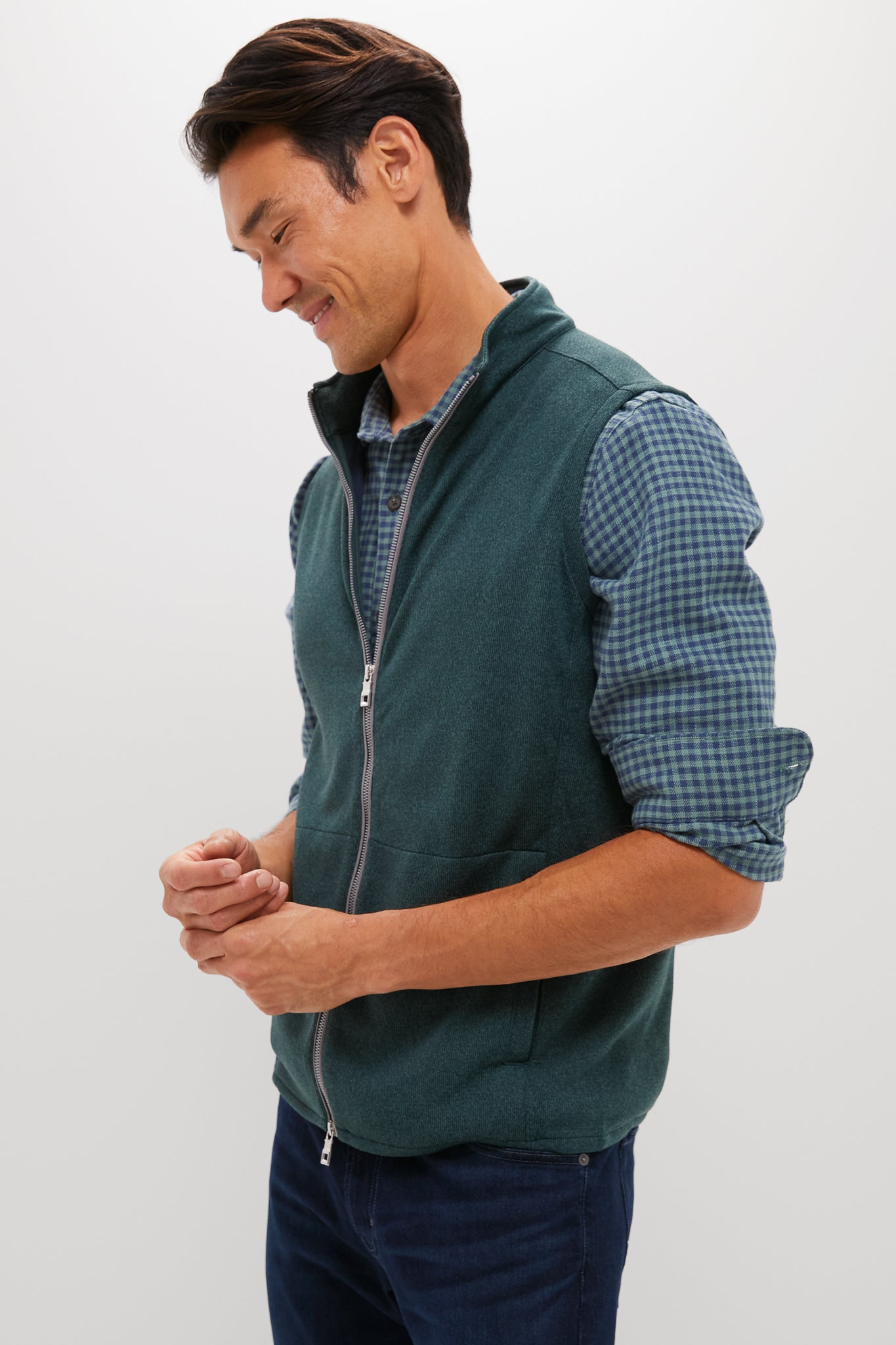 A man in a green Peter Millar quarter-zip sweater over a blue plaid shirt stands looking down and smiling, hands loosely clasped.