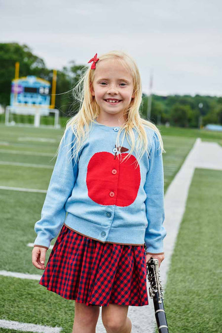 Young girl on a soccer field wearing the classic Light Blue Apple Constance Sweater with a red apple motif, showcasing effortless, fun style, holding a clarinet.