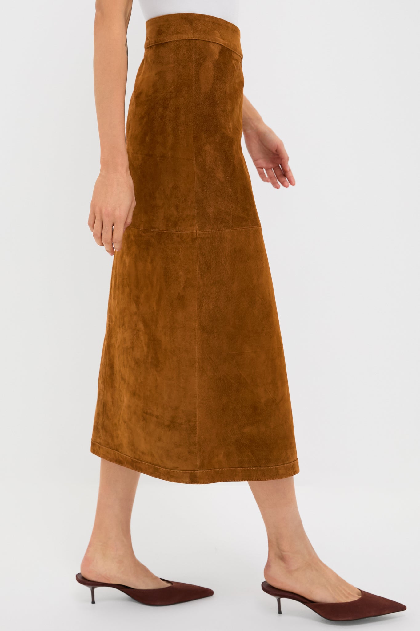 Model wearing Nutmeg Winona Skirt—a classic, suede A-line midi with a wrap design and snap closure, paired effortlessly with matching mules and a white top.