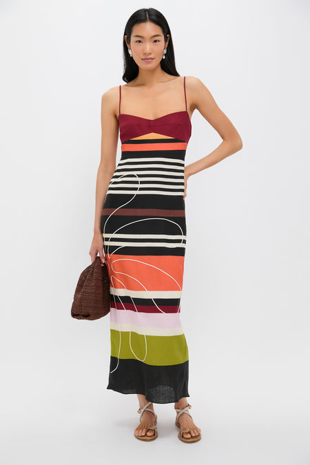 A woman in a Cala de la Cruz colorful striped midi dress with an empire waistline and cutout detail holds a brown woven bag and wears sandals, standing against a plain background.