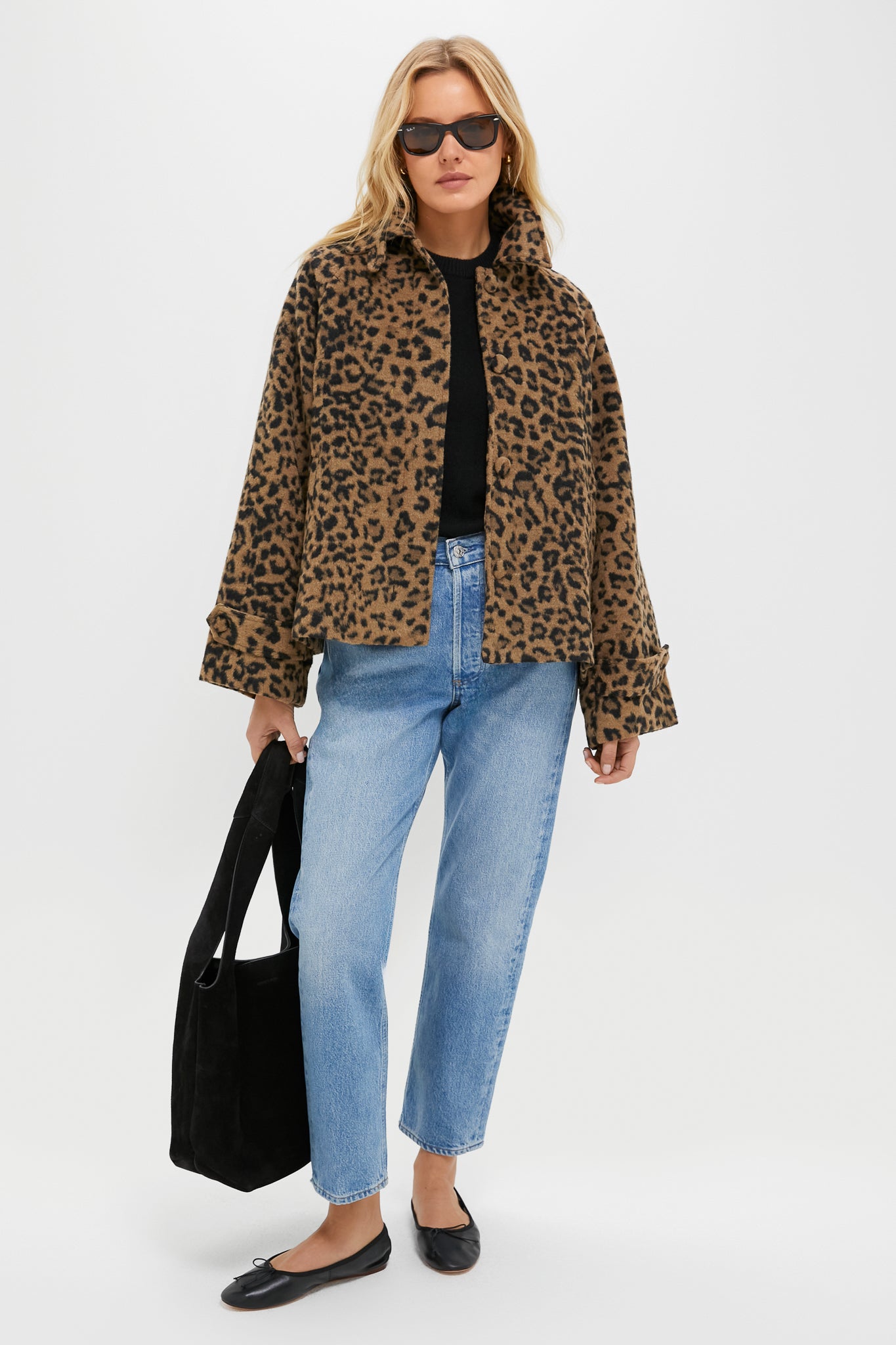 A woman wears a Tuckernuck leopard print oversized jacket, black top, light blue jeans, black flats, sunglasses, and carries a large black tote bag while standing against a white background.