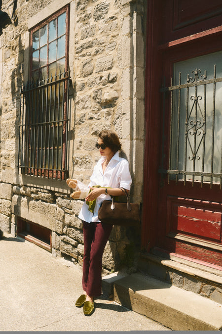 Woman leaning on a building in a white button down, burgundy pants, green loafers, and a brown suede bag. 