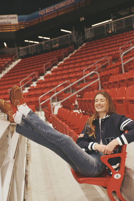 Young woman lounges with a smile in a mostly empty stadium, wearing Overcast High Rise Straight 90's Pinch Waist Long jeans, exuding a classic, effortless, and high-end style.