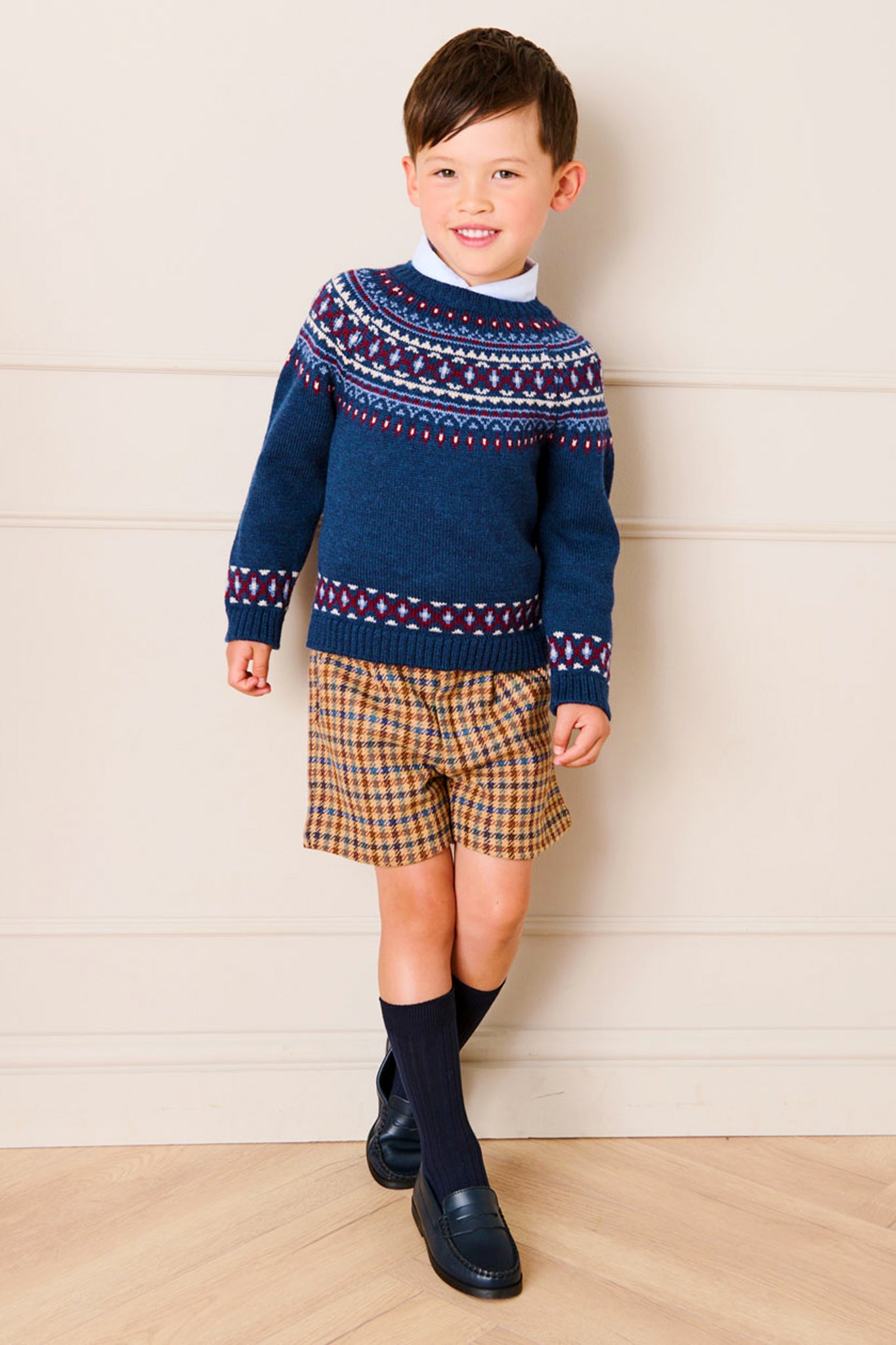 A young boy stands indoors, smiling, dressed in a Pepa London blue Fair Isle jacquard sweater, plaid shorts, navy knee socks, and black shoes—a charming showcase of classic children's fashion.
