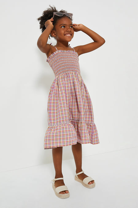 A young girl wearing a Bebe Organic pink plaid seersucker cotton smocked summer dress with white sandals and sunglasses highlights stylish organic children's wear.