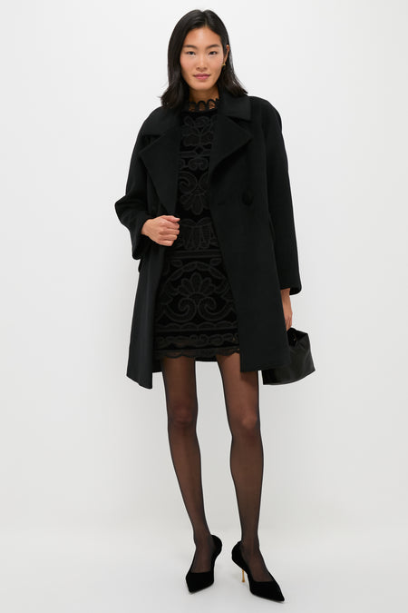 A woman stands against a white background wearing Tuckernuck’s double-breasted black coat over a patterned black dress, sheer tights, black heels, and carrying a small black handbag.