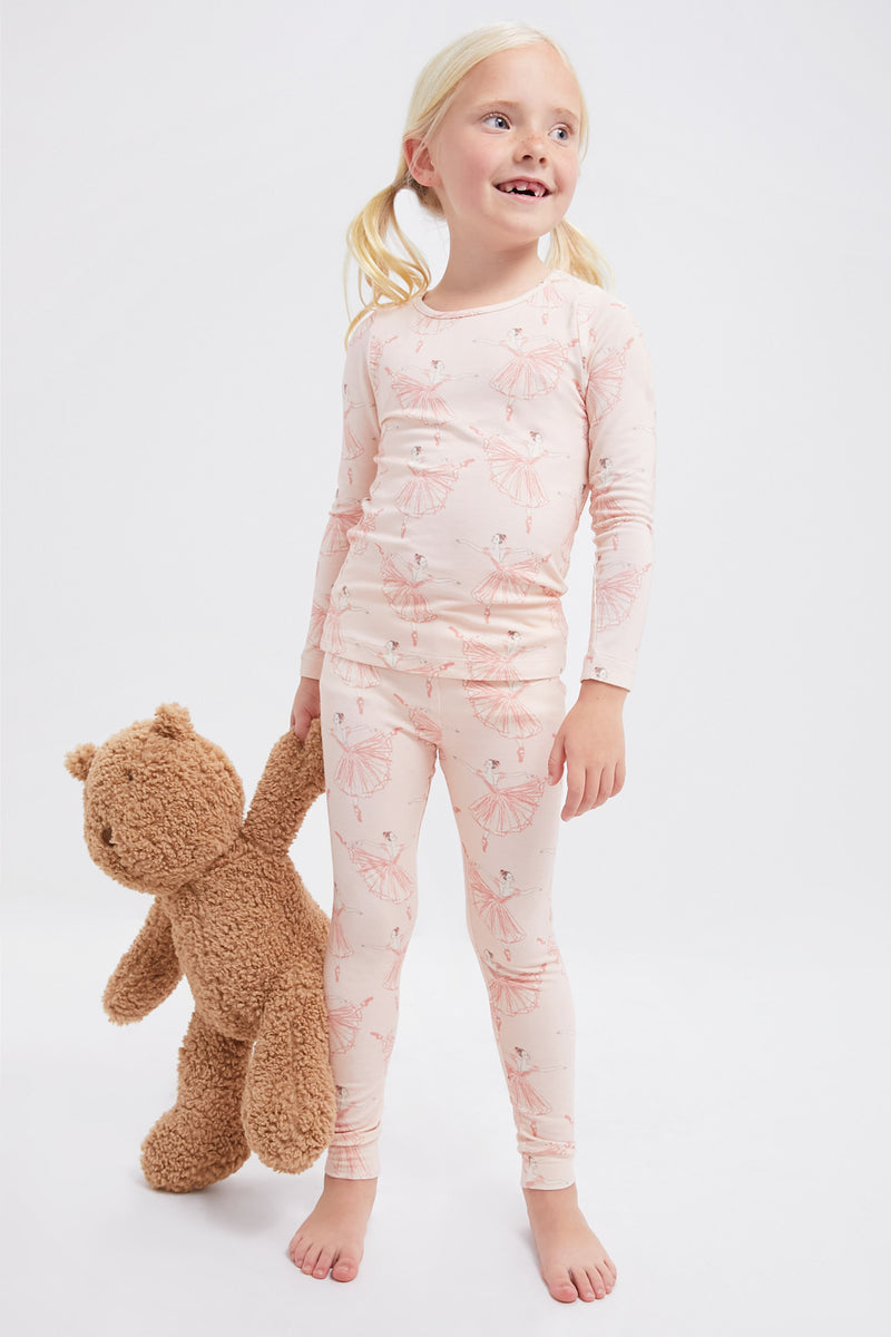 A young girl in Pink Chicken kids’ pajamas with umbrella print stands barefoot, holding a brown teddy bear by the arm against a plain white background.