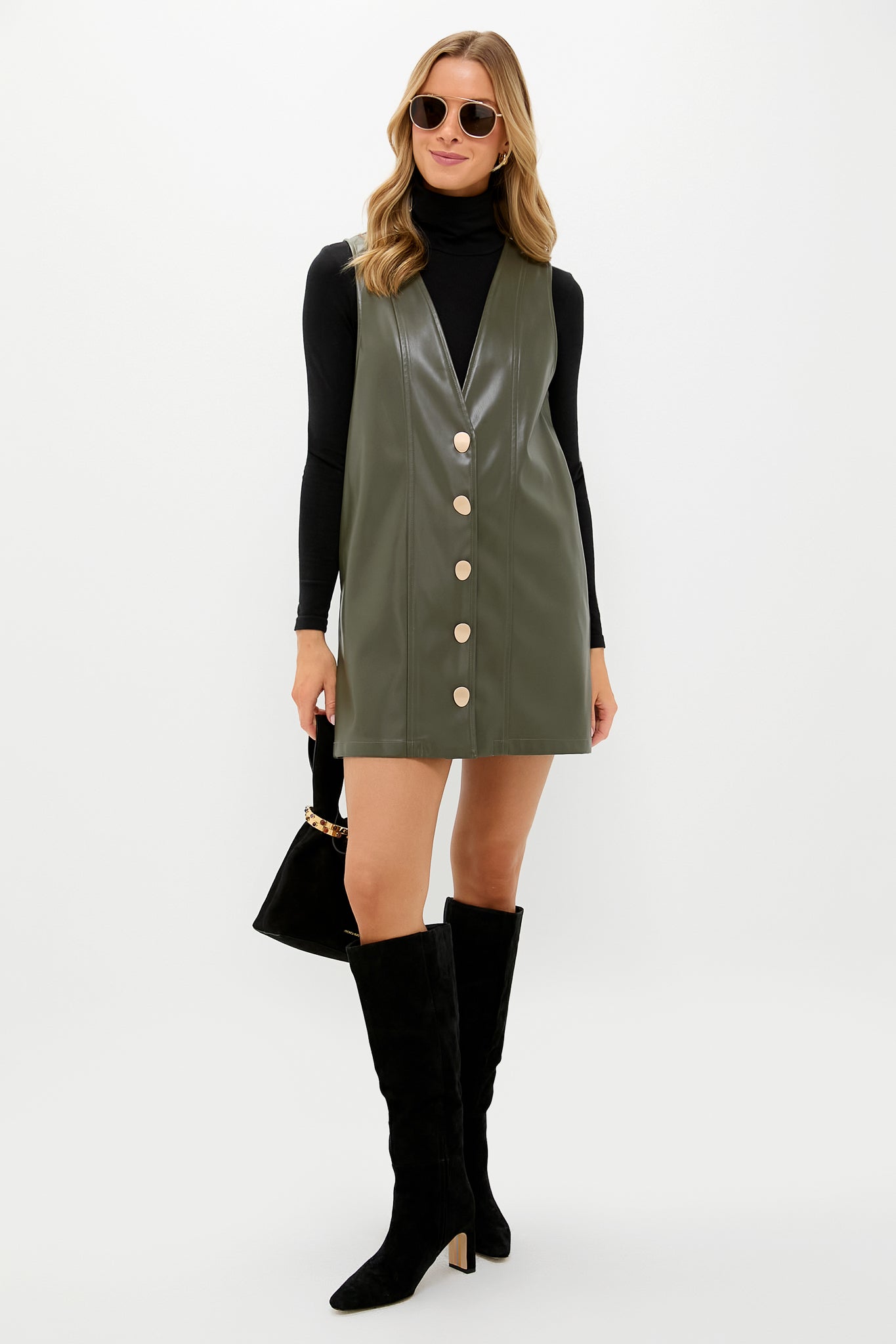 Model in Olive Faux Leather Pinafore Dress over a fitted black turtleneck, styled with suede knee-high boots, gold accessories, and a chic black handbag for an effortless, classic look.