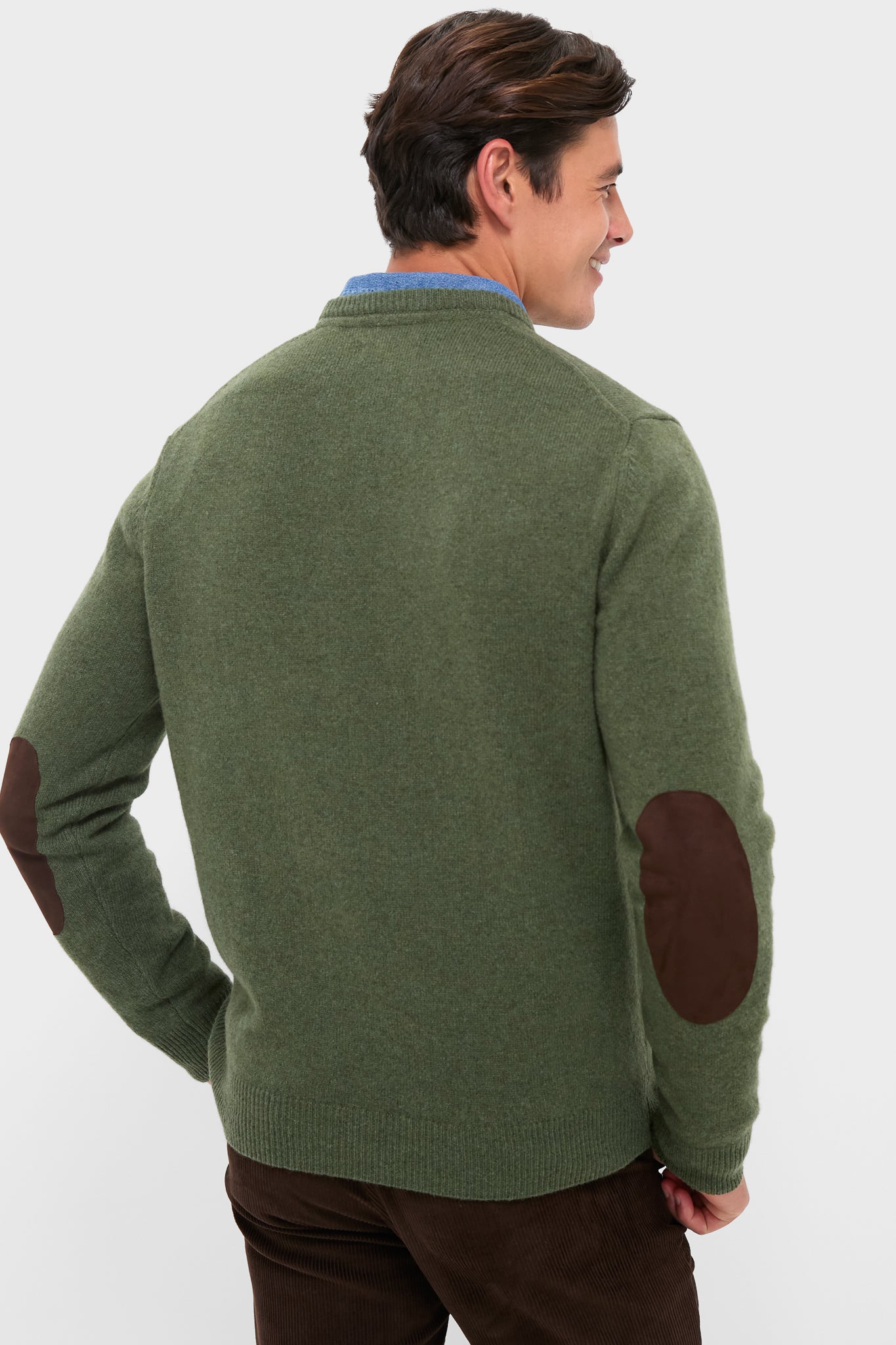 Man wearing classic Olive Essential Patch Crew Neck Sweater with leather elbow patches, blue shirt, and corduroy trousers, exuding effortless, high-end style in a studio setting.