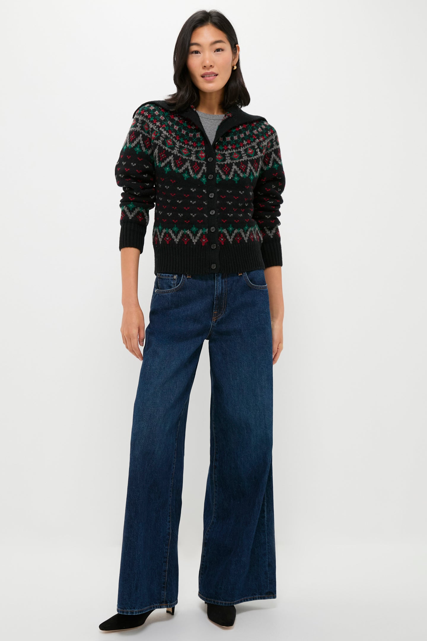 A woman stands against a plain background wearing a Polo Ralph Lauren Fair Isle cardigan, a gray shirt, wide-leg blue jeans, and black shoes.