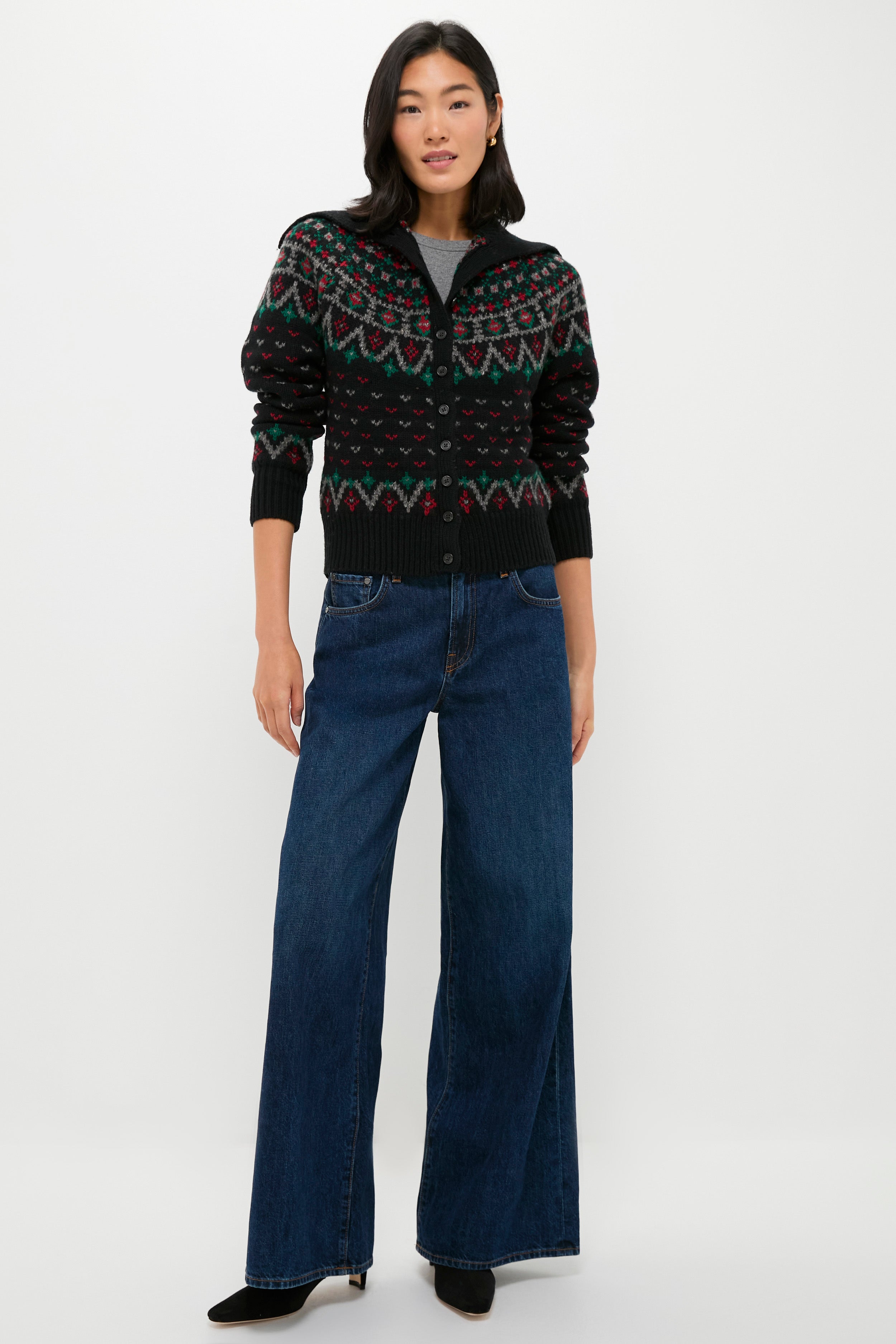 A woman stands against a plain background wearing a Polo Ralph Lauren Fair Isle cardigan, a gray shirt, wide-leg blue jeans, and black shoes.