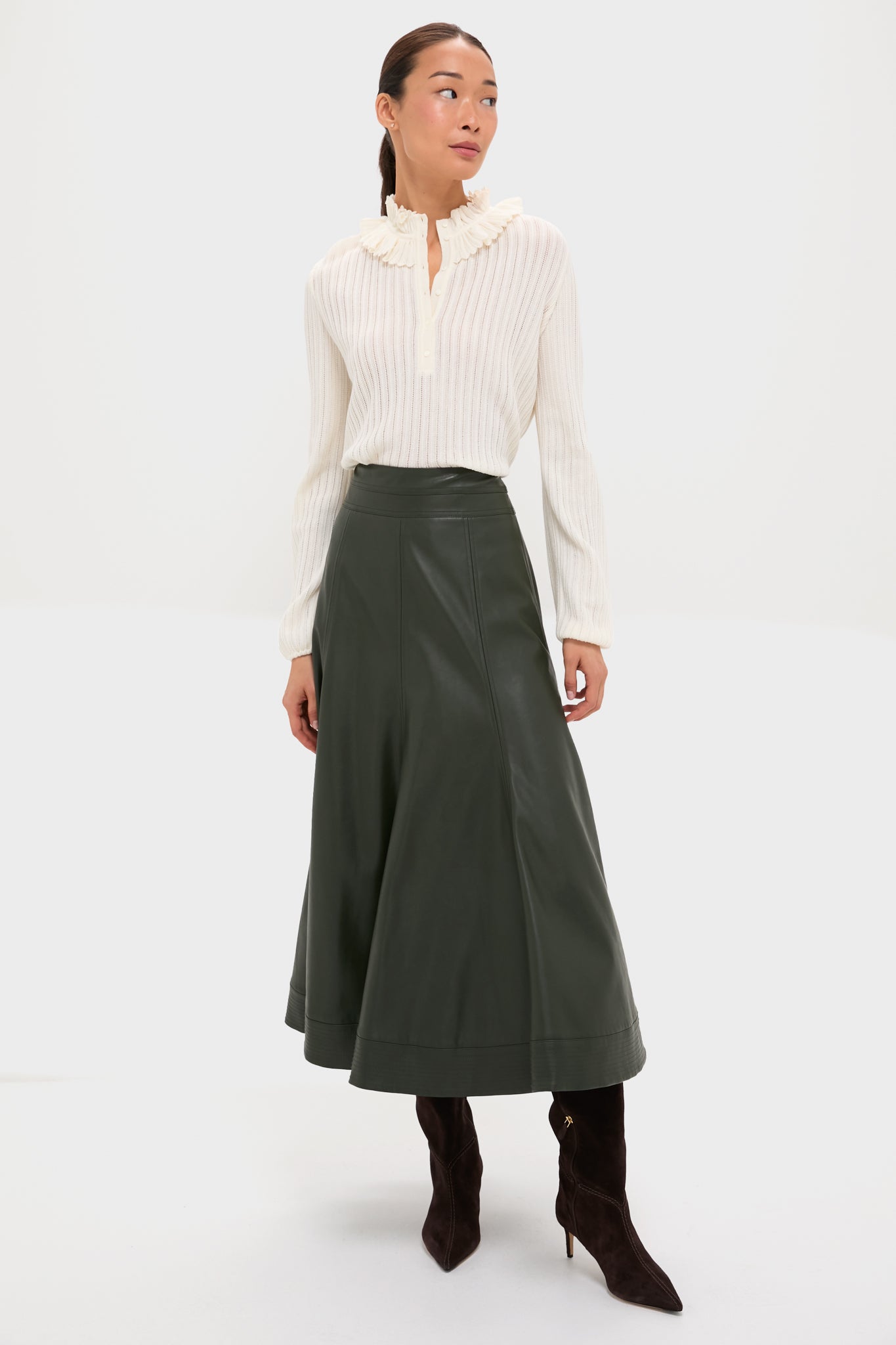 A woman in a molli cream openwork knit blouse, dark green midi leather skirt, and dark brown knee-high boots stands against a plain white background.