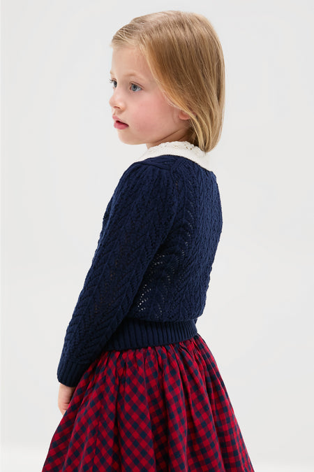 A young child with blond hair stands against a light background, wearing a Polo Ralph Lauren navy blue baby sweater and red plaid skirt, looking to the side.