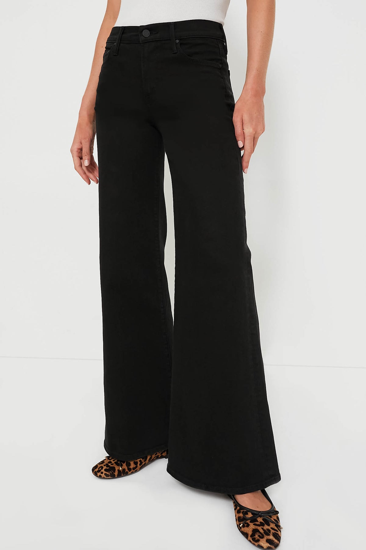 A person wears MOTHER high-waisted black wide-leg jeans paired with a white ribbed sleeveless top and leopard print flats. The black denim from MOTHER features a stylish wide leg for a modern vibe.