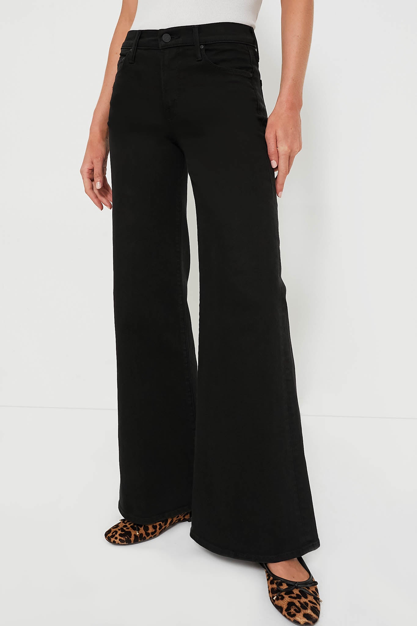 A person wears MOTHER high-waisted black wide-leg jeans paired with a white ribbed sleeveless top and leopard print flats. The black denim from MOTHER features a stylish wide leg for a modern vibe.