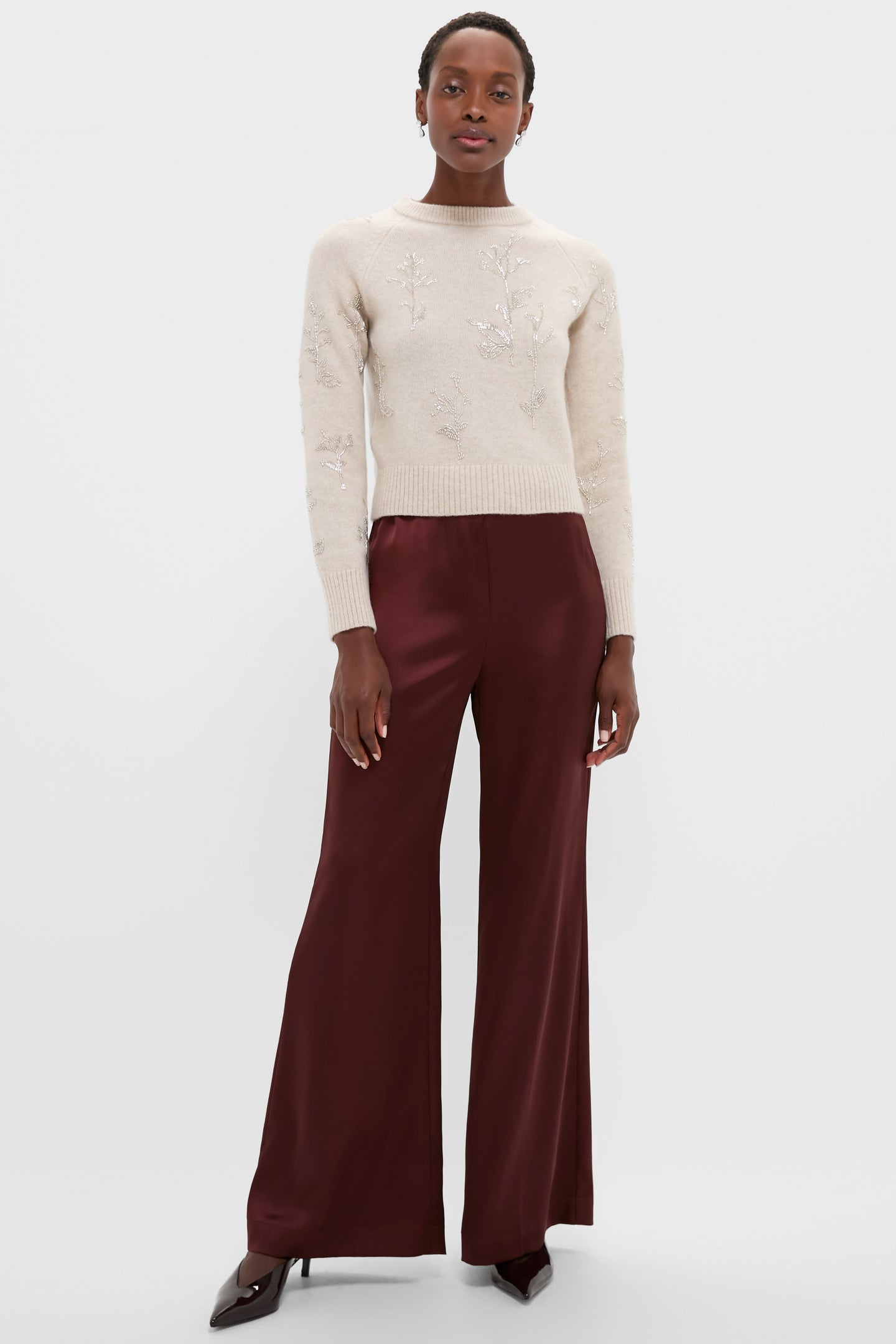 A person in an Altuzarra cream crewneck sweater with delicate beadwork, burgundy satin wide-leg pants, and dark pointed-toe heels stands against a plain background.