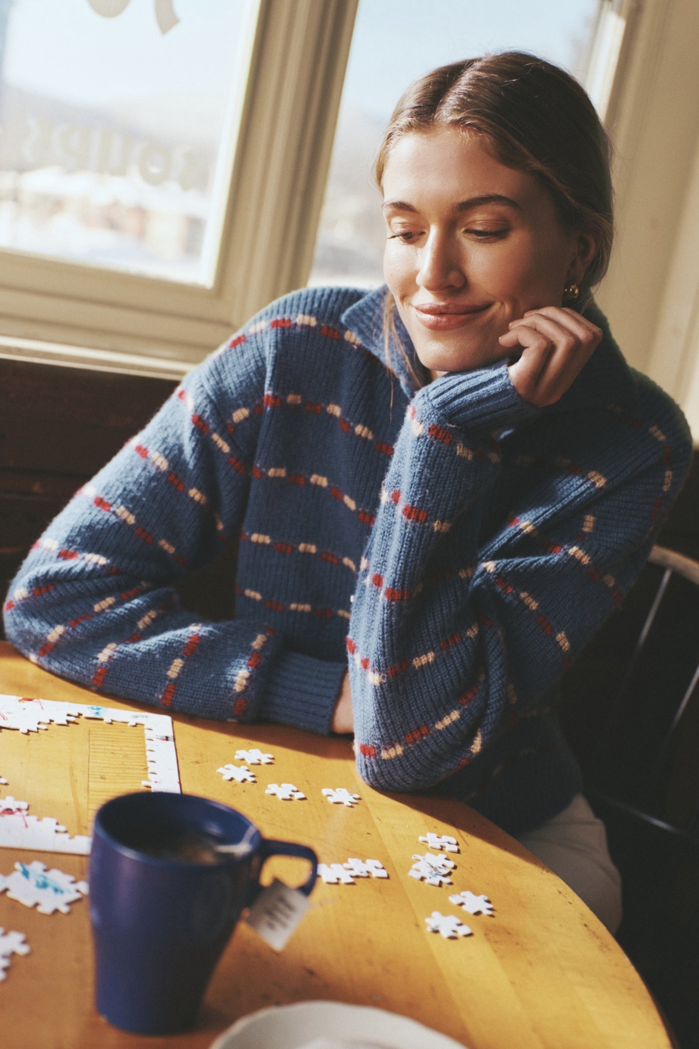 Young woman in a classic Navy Striped Jansen V-Neck Polo Sweater, thoughtfully assembling a puzzle at a café table, embodying effortless and high-end style.