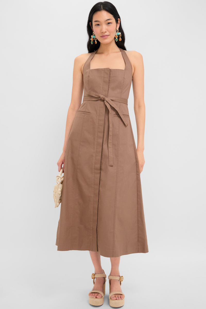 A woman stands against a plain background wearing a sleeveless brown Cult Gaia halter-neck midi dress with a belt, platform sandals, colorful earrings, and holding a small bag.