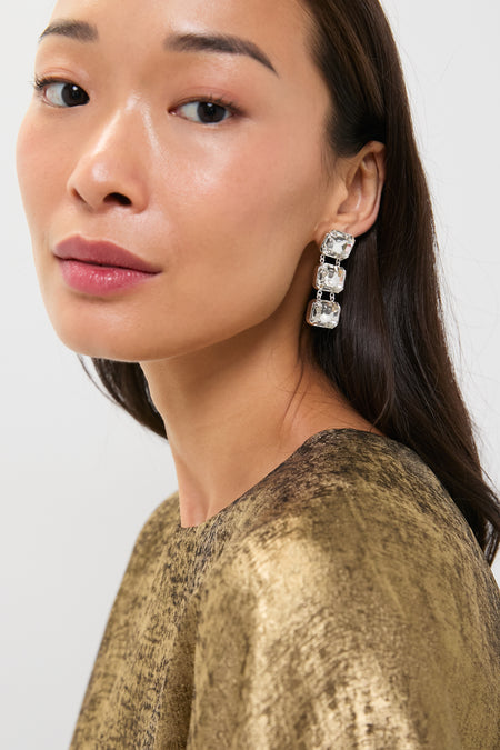 A woman with long dark hair wears a metallic gold top and striking Tuckernuck Jewelry gold plated brass dangle earrings with glass crystals, looking over her shoulder against a plain background.