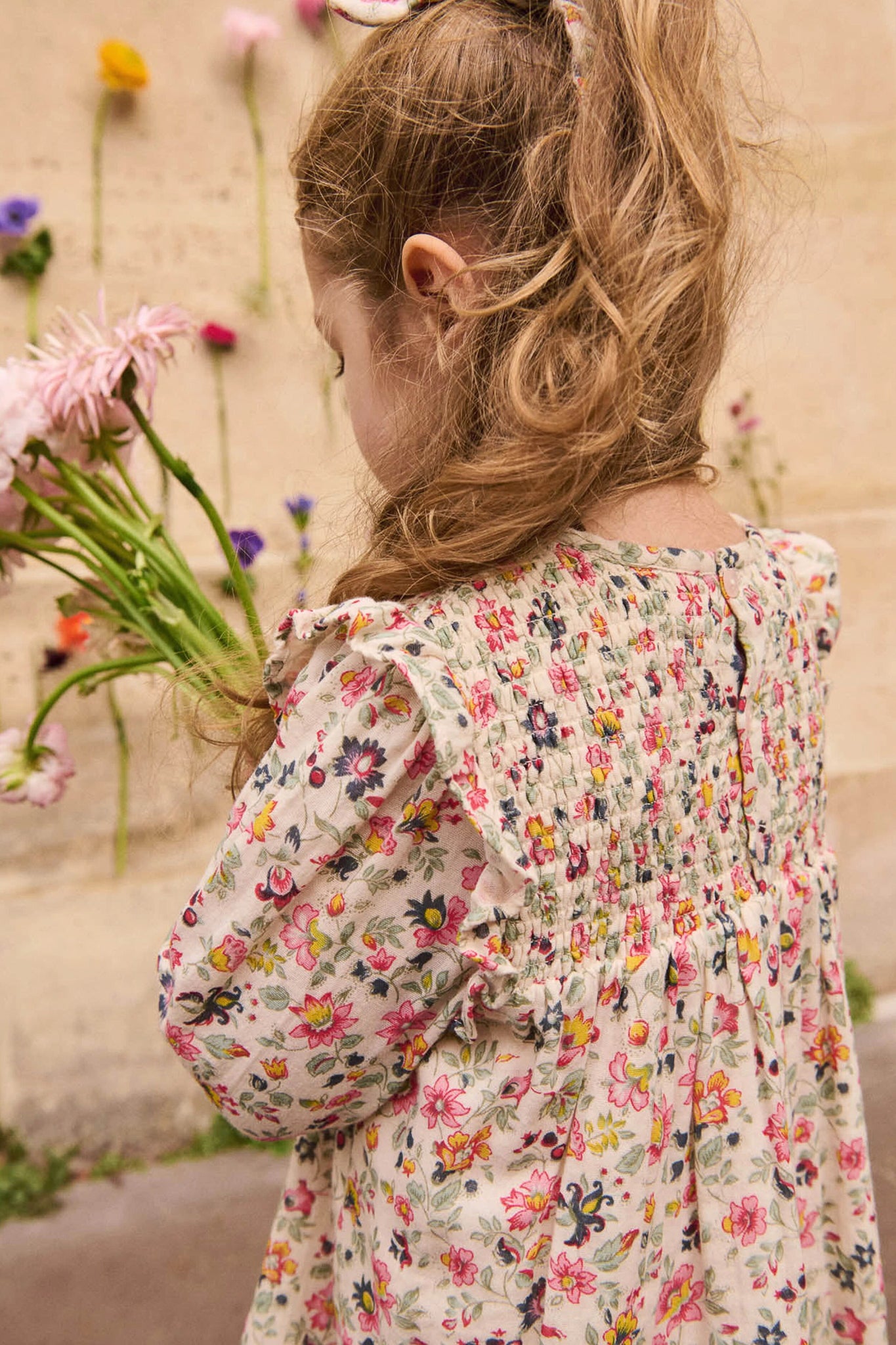 Young girl in a classic Cream Wild Flowers Saralou Dress, effortlessly holding a bouquet, showcasing fun, high-end floral print with ruffles and smocked bodice.