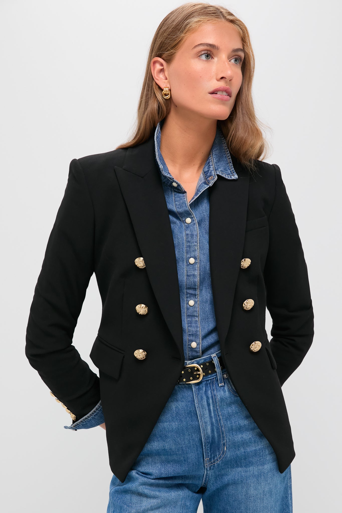 A woman wears a black blazer with gold buttons from Veronica Beard over a denim shirt and jeans, standing with her hands in her pockets.