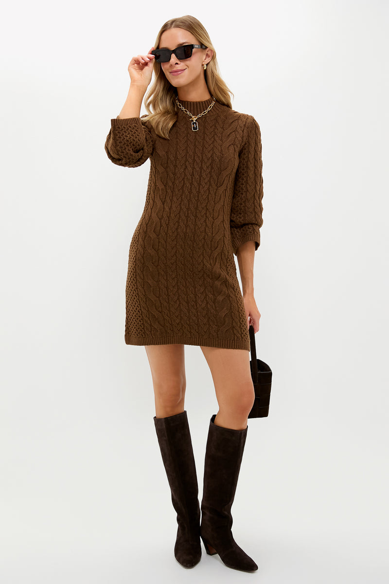 A woman wearing a Loeffler Randall brown cable-knit sweater dress, knee-high dark brown boots, black handbag, necklace, and black sunglasses stands against a white background.