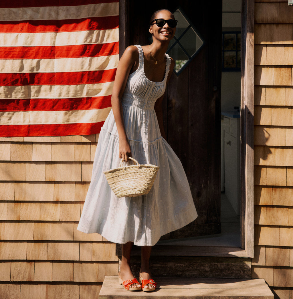 Woman standing in front of a door in a blue gauze dress and red heels holding a straw bag. 