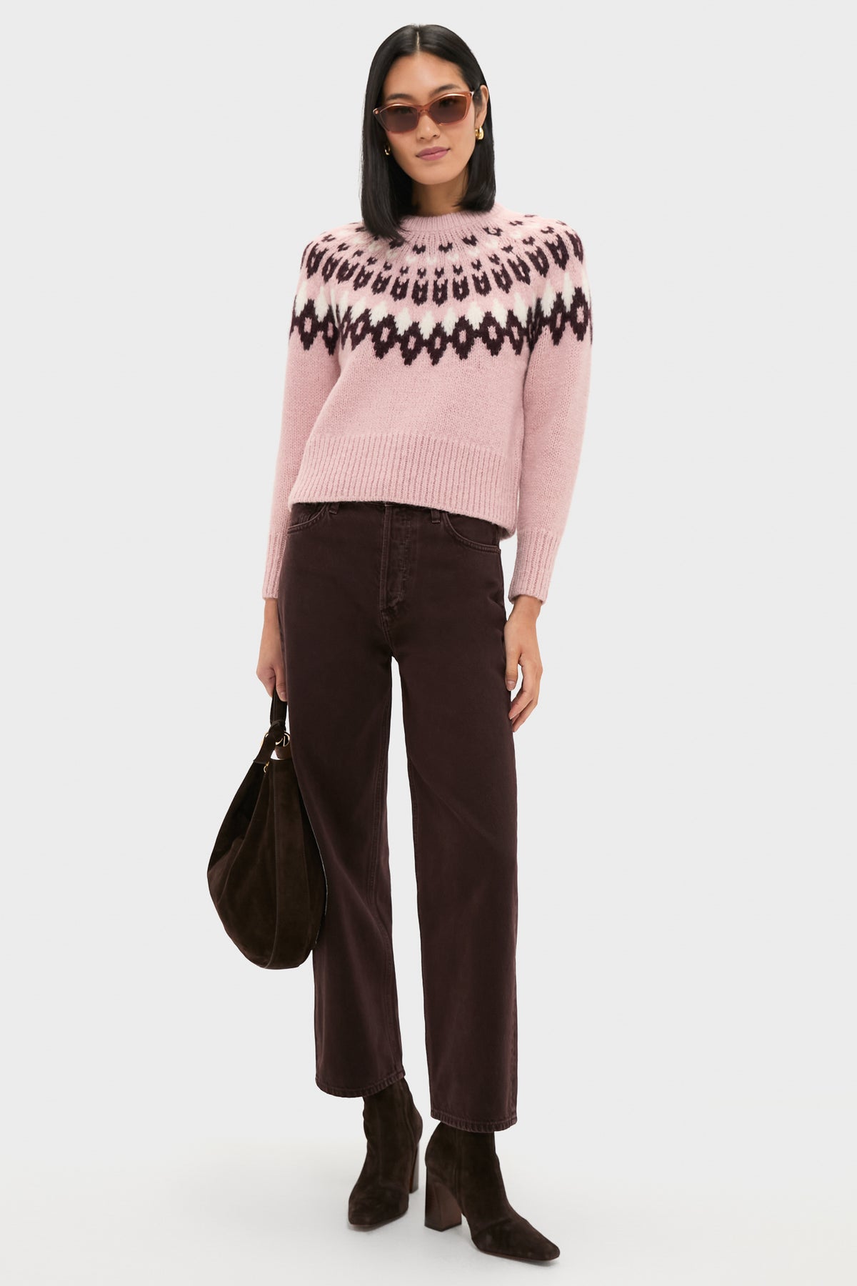 A woman wearing a pink Fair Isle sweater by Veronica Beard, paired with brown wide-leg pants, dark boots, sunglasses, and holding a brown bag, stands against a plain background.