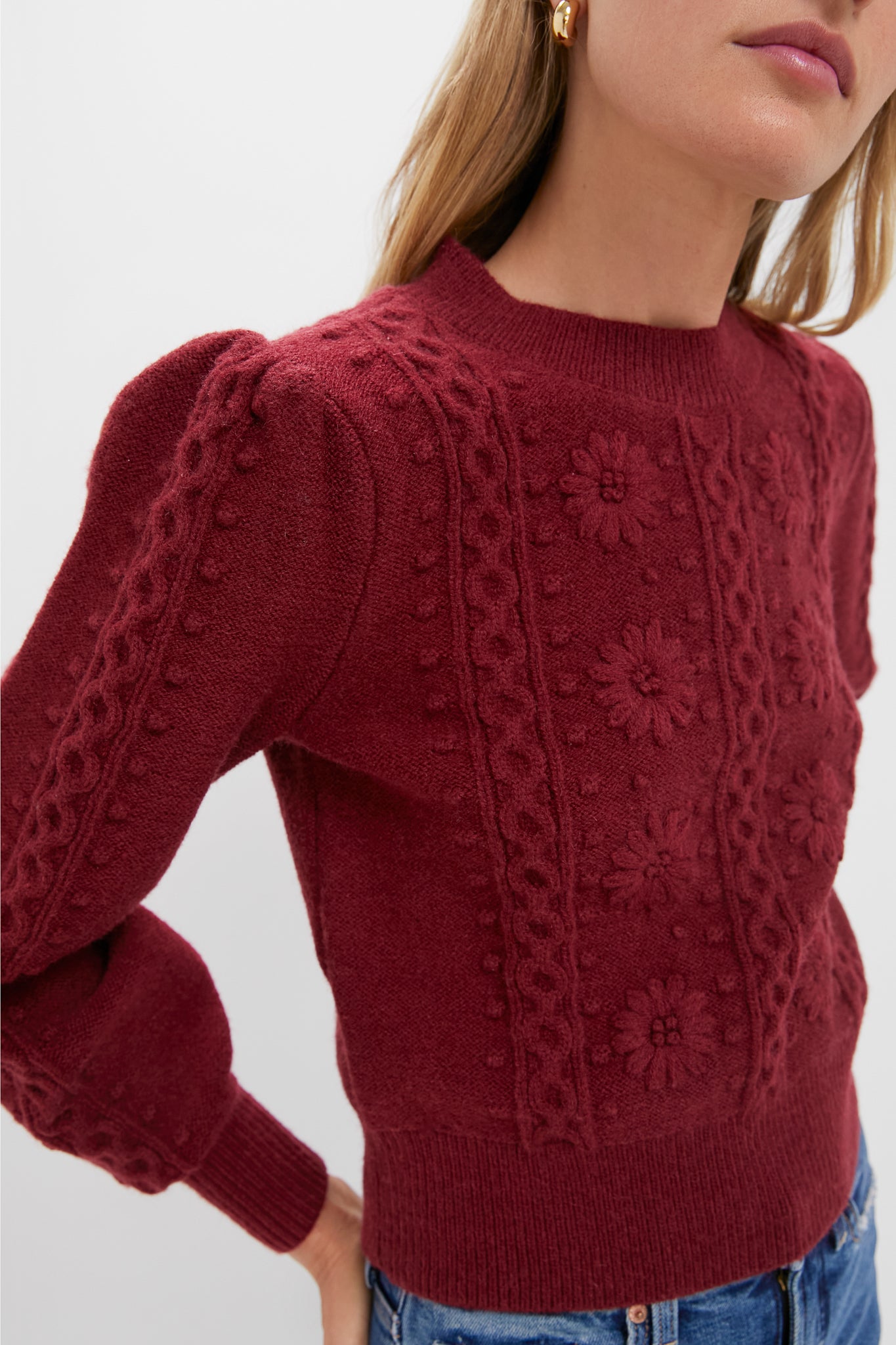Person wearing the Merlot Embroidered Floral Frankie Sweater with cable-knit and bobble-stitch detailing, offering a classic and effortless look, paired with blue jeans.