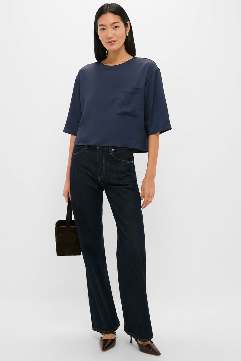 A woman wears a TWP navy boxy blouse with a chest pocket, dark straight-leg jeans, pointed-toe heels, and holds a brown handbag against a plain white background.