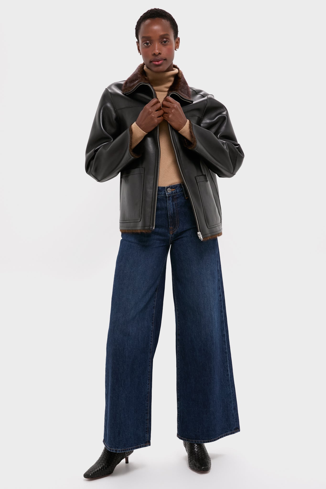 Wearing a reversible black faux leather jacket with brown collar by Stand Studio, a beige turtleneck sweater, wide-leg blue jeans, and black ankle boots, a person poses against a white background. The versatile jacket elevates the stylish ensemble.