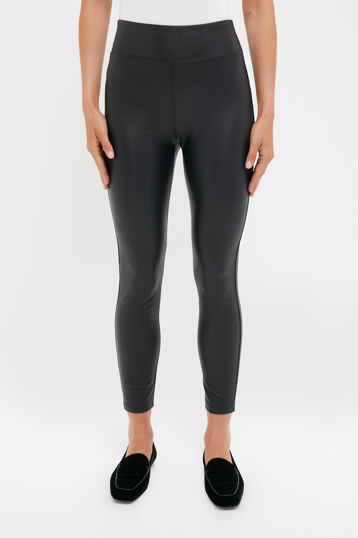 A person wearing Tuckernuck high waisted black faux leather leggings and black loafers stands against a plain white background.