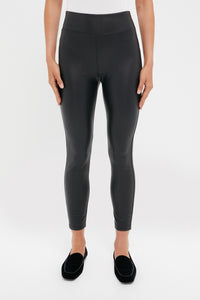 A person wearing Tuckernuck high waisted black faux leather leggings and black loafers stands against a plain white background.