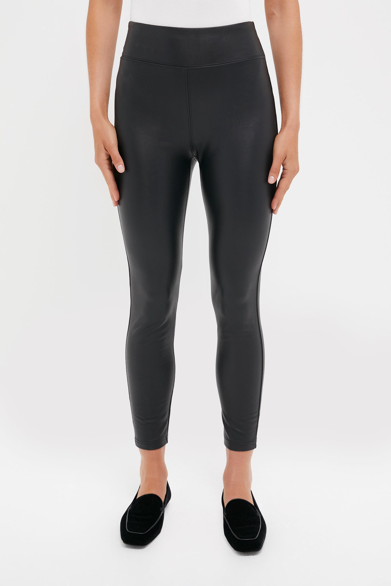 A person wearing Tuckernuck high waisted black faux leather leggings and black loafers stands against a plain white background.