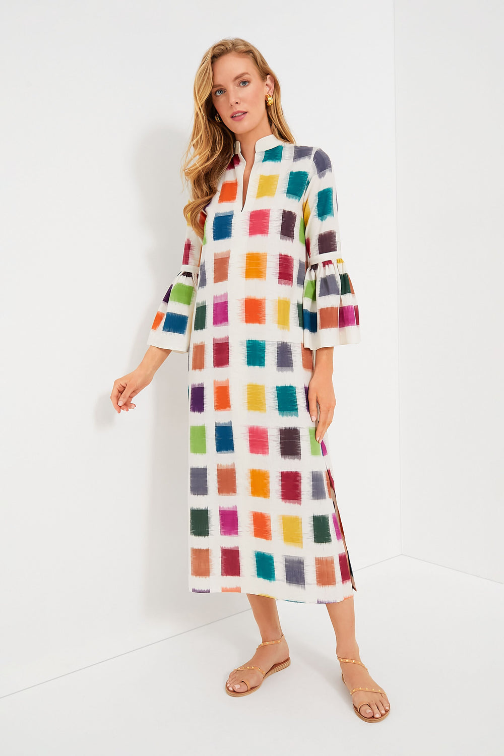 A woman wears a De Castro Rainbow Parampara Dress—an artisanal, long white organic cotton dress with a vibrant square pattern and three-quarter bell sleeves—paired with beige sandals.