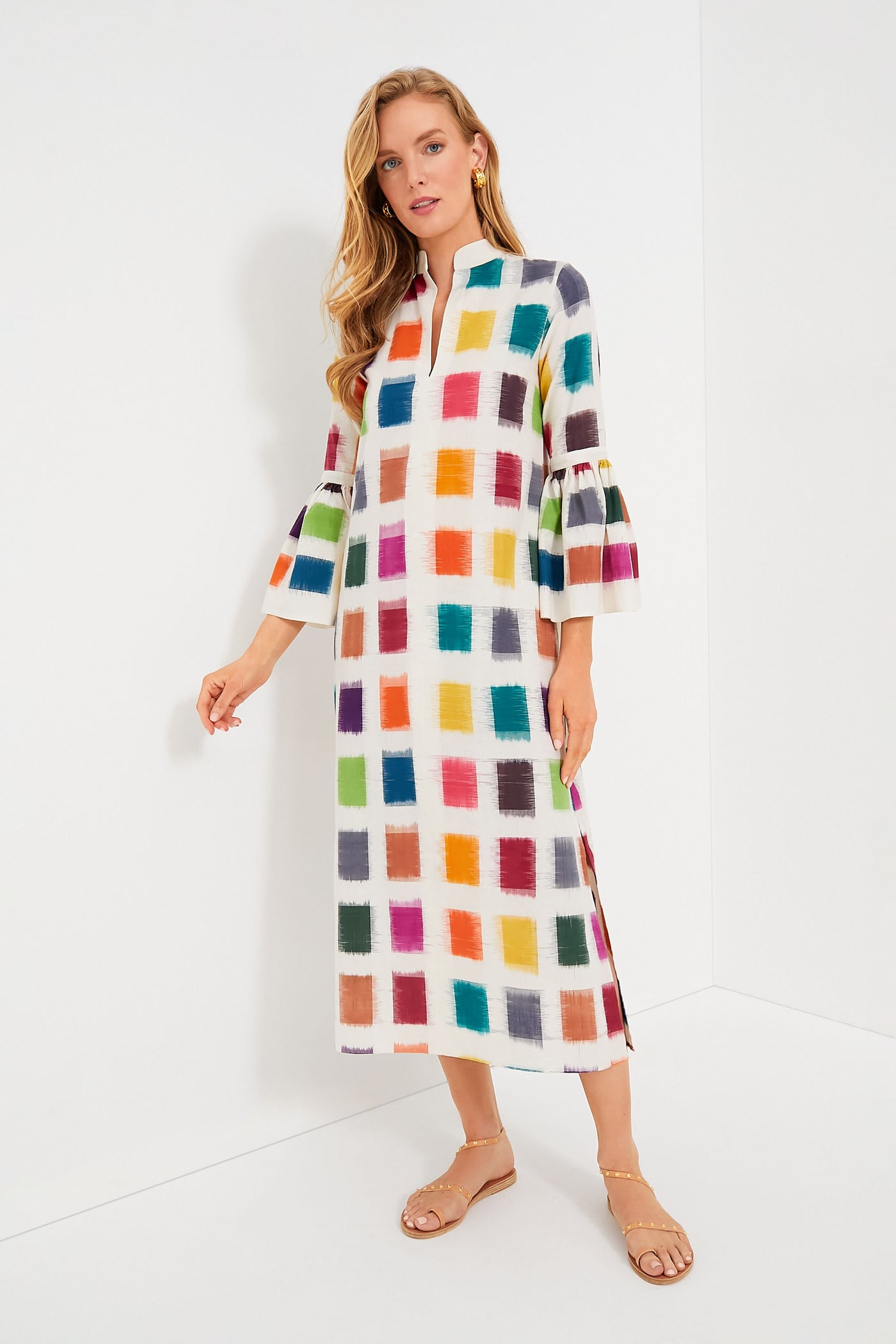 A woman wears a De Castro Rainbow Parampara Dress—an artisanal, long white organic cotton dress with a vibrant square pattern and three-quarter bell sleeves—paired with beige sandals.