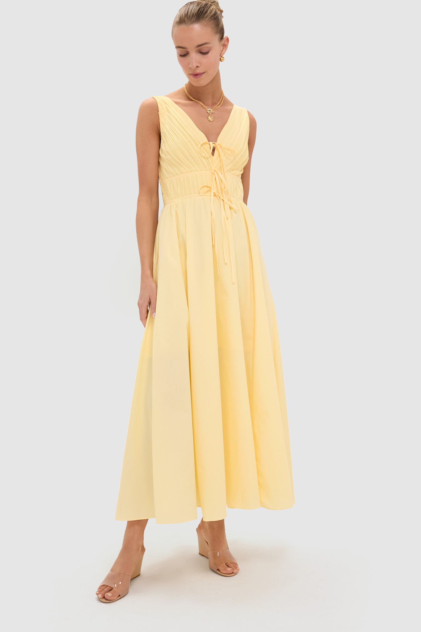 A woman wears a Moon River sleeveless light yellow maxi dress with a fitted bodice, cinched waist, and tie detail, styled with clear heeled sandals—an ideal breezy summer outfit.